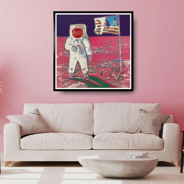 Framed artwork of an astronaut on the moon with a flag, displayed above a white sofa in a room with pink walls.