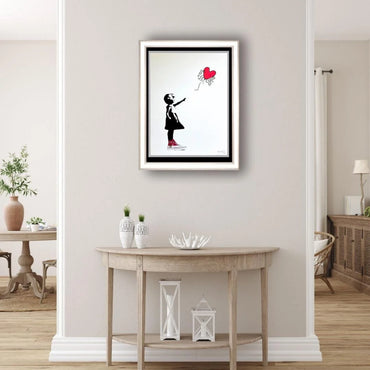 Framed artwork of a girl with a red balloon on a wall above a wooden table.